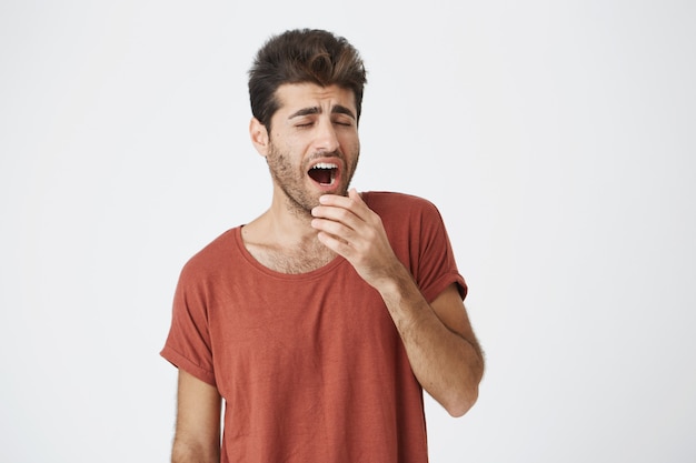 Gapende jonge kerel met baard en trendy kapsel moe van het werk en hield zijn hand achter zijn mond. Studenten die een rood T-shirt droegen, raakten de lezingen zat