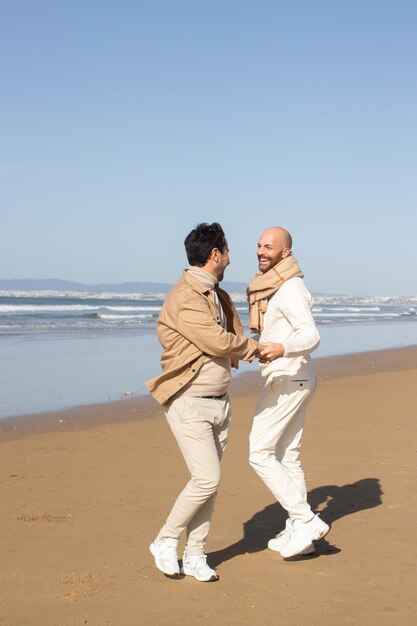 Gay paar ronddraaien en hand in hand op het strand. Vrolijke opgewonden mannen van middelbare leeftijd die elkaar aankijken tijdens het dansen op zand. Datingconcept