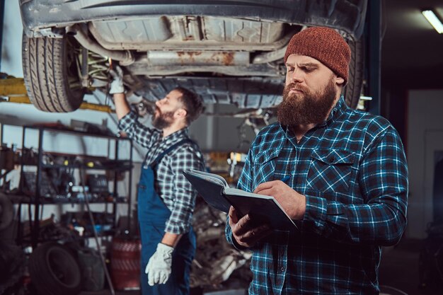 Gebaarde deskundige monteur registreert resultaten van het inspecteren van een auto terwijl zijn partner een probleem met een auto-ophanging controleert. Een scène uit een tankstation.