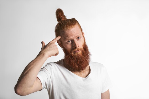 Gebruik je hersens. Portret van emotionele verontwaardigde jeugd mannelijke hipster met bijgesneden stijlvolle baard en haar knoop poseren bij witte studio muur, wijsvinger draaien op zijn tempel, met perplex blik