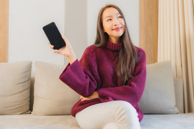 Gebruik slimme mobiele telefoon van de portret mooie jonge Aziatische vrouw op bank in woonkamerbinnenland