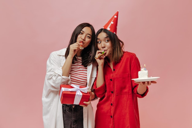 Gebruinde Aziatische vrouwen blazen partij hoorns op geïsoleerde Leuke vrouw in rode blouse en feestmuts houdt verjaardagstaart Jonge dame in stijlvolle outfit poses met geschenkdoos op roze achtergrond