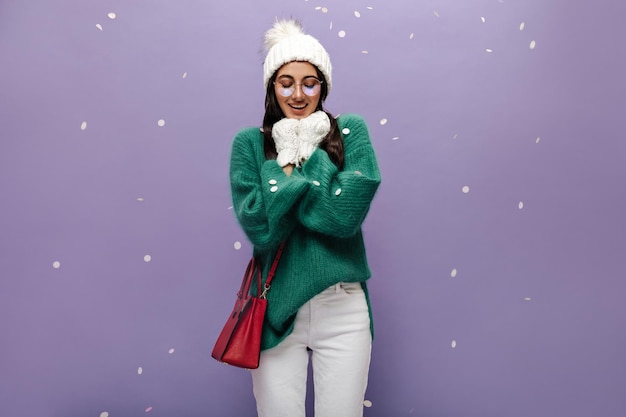 Gebruinde brunette vrouw in brillen gebreide muts en handschoenen warmt op en glimlacht op paarse achtergrond met sneeuwvlokken