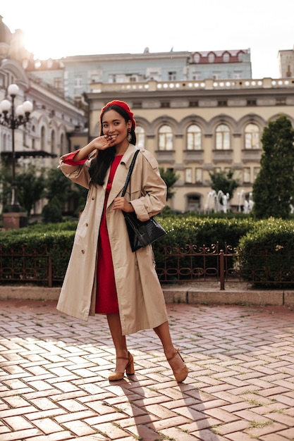 Gebruinde brunette vrouw in midi rode jurk, stijlvolle baret en beige trenchcoat lacht breed en poseert buiten