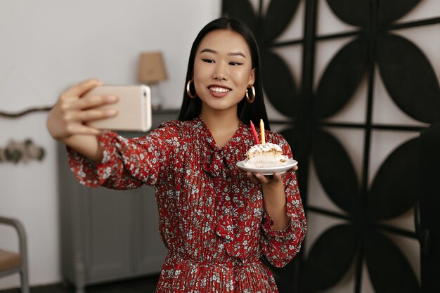 Gebruinde brunette vrouw in rode bloemenjurk houdt telefoon vast en neemt selfie met lekkere stukjes verjaardagstaart