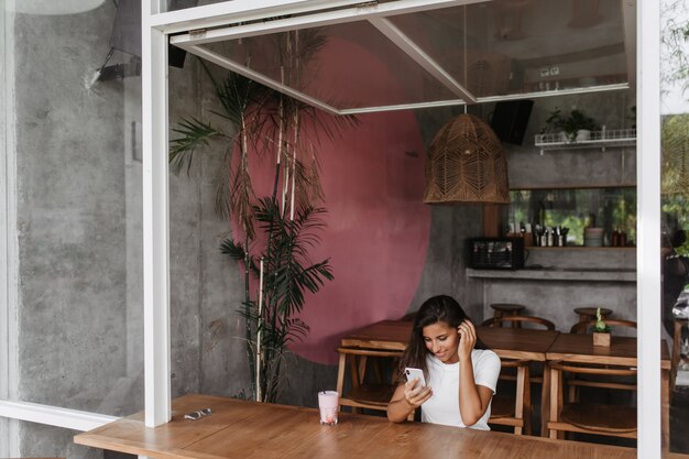 Gebruinde dame zit in café met houten meubilair en kijkt naar video op telefoon