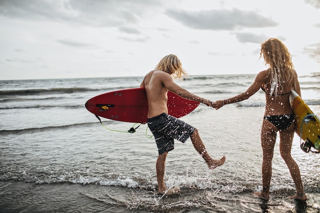 Gebruinde man en vrouw houden hand vast, houden surfplanken vast en spatten water