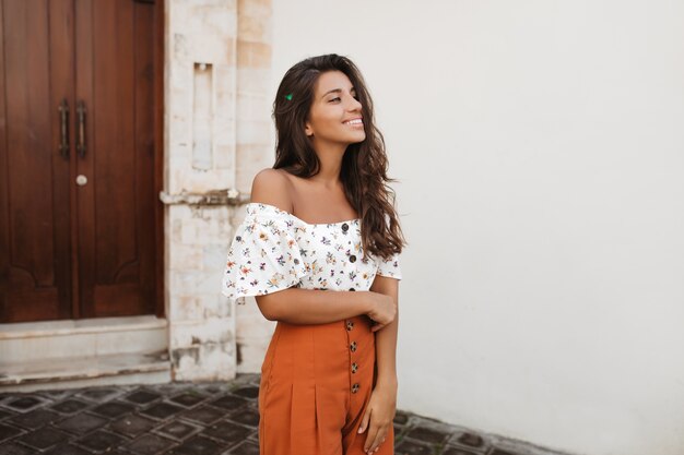 Gebruinde vrouw in stijlvolle oranje korte broek met hoge taille en lichte blouse poseren tegen muur van huis met antieke houten deuren