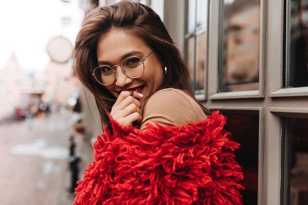 Gebruinde vrouw met een stijlvolle bril lacht voor haar mond Portret van een meisje met rode lippenstift gekleed in een trendy oversized trui