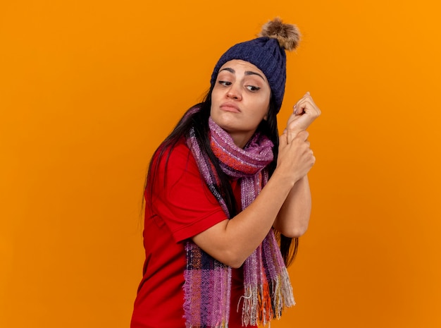 Geconcentreerd jong Kaukasisch ziek meisje dat de winterhoed en sjaal draagt die impuls controleert die kant bekijkt die op oranje muur met exemplaarruimte wordt geïsoleerd