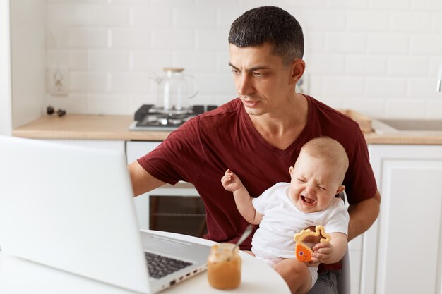 Geconcentreerde aantrekkelijke brunette man freelancer dragen casual stijl kastanjebruin t-shirt, werken op laptop en zorgen voor zijn dochtertje, zittend in witte keuken.