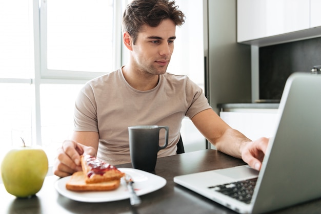 Geconcentreerde aantrekkelijke mens die laptop met behulp van terwijl het eten van ontbijt