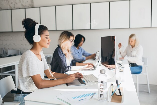 Geconcentreerde Afrikaanse vrouwelijke webdesigner met grafisch tablet terwijl haar collega's rapporten schrijven. Indoor portret van programmeurs van een internationaal bedrijf dat samen tijd doorbrengt op de werkplek.