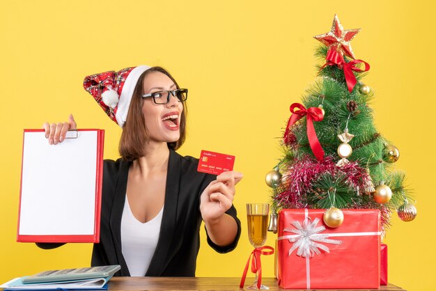 Geconcentreerde charmante dame in pak met kerstman hoed en bril met bankkaart en document in het kantoor op geel geïsoleerd