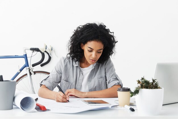 Geconcentreerde gemengde rasvrouw met zwart krullend haar dat op haar werkplaats is