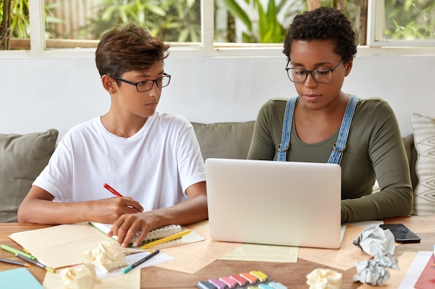 Geconcentreerde hipster schrijft in notitieboekje informatie die hij hoort van een vrouw die nieuws leest van de internetwebsite. Mooie zwarte meisjestoetsenborden bij laptopcomputer