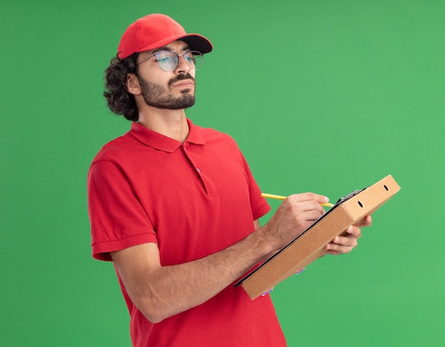 Geconcentreerde jonge blanke bezorger in rood uniform en pet met bril met pizzapakket schrijvend op klembord met potlood kijkend naar klembord