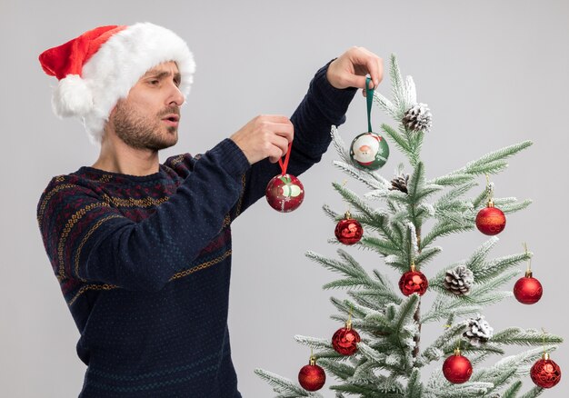 Geconcentreerde jonge blanke man met kerstmuts staande in de buurt van de kerstboom en versiert het met kerst ornament ballen geïsoleerd op een witte achtergrond