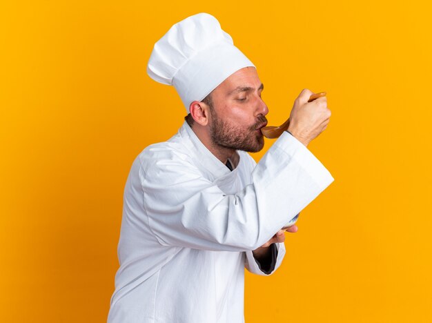 Geconcentreerde jonge blanke mannelijke kok in uniform van de chef-kok en pet staande in profielweergave met kom testmaaltijd met lepel geïsoleerd op oranje muur