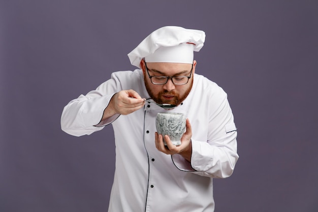 Geconcentreerde jonge chef-kok met een uniforme bril en een pet die een kom vasthoudt alsof hij uit een kom eet met een lepel die naar beneden kijkt geïsoleerd op een paarse achtergrond
