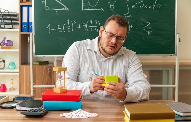 Geconcentreerde jonge leraar met een bril die aan het bureau zit met schoolbenodigdheden in de klas die met pen op aantekeningen op papier schrijft