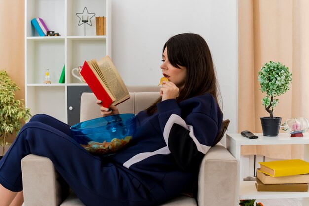 Geconcentreerde jonge vrij Kaukasische vrouw zittend op een fauteuil in ontworpen woonkamer met kom met chips op benen aardappelchip eten en boek lezen