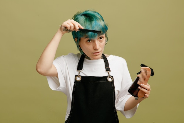 Geconcentreerde jonge vrouwelijke kapper in uniform met haarlak en kam die haar eigen haar kamt en omhoog kijkt geïsoleerd op olijfgroene achtergrond