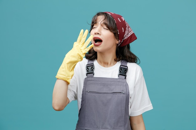 Geconcentreerde jonge vrouwelijke reiniger met uniforme bandana en rubberen handschoenen die de hand voor de mond houden niezen met gesloten ogen geïsoleerd op blauwe achtergrond