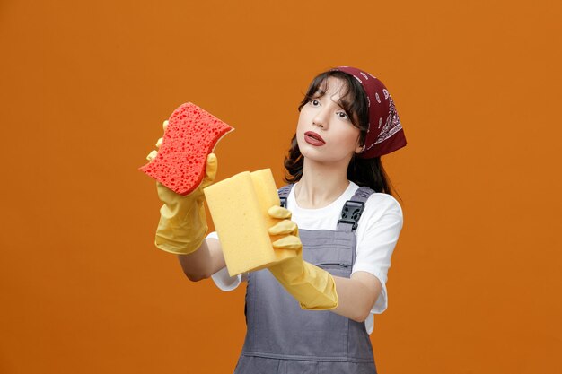 Geconcentreerde jonge vrouwelijke reiniger met uniforme rubberen handschoenen en bandana die sponzen uitrekt en naar de zijkant kijkt alsof ze iets schoonmaakt dat geïsoleerd is op een oranje achtergrond