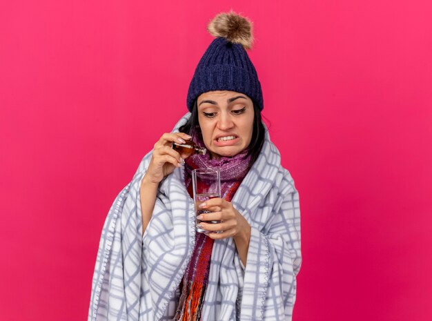Geconcentreerde jonge zieke vrouw die de wintermuts en sjaal draagt die in plaid wordt verpakt die geneesmiddel toevoegt aan glas water dat op roze muur wordt geïsoleerd