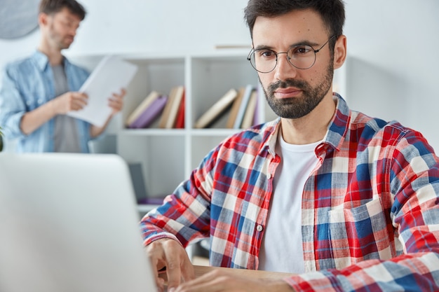Geconcentreerde man freelancer werkt in de verte op laptop, heeft stoppels en draagt een bril