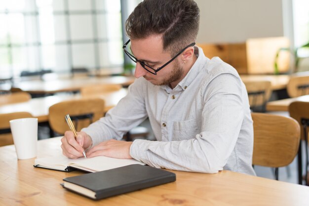 Geconcentreerde mannelijke student die in notitieboekje bij bureau in klaslokaal schrijft