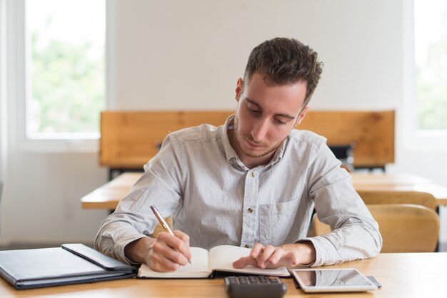 Geconcentreerde mannelijke student die thuiswerk doen bij bureau in klaslokaal