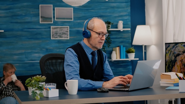 Geconcentreerde oude ondernemer met koptelefoon die naar muziek luistert terwijl hij afbeeldingen controleert die schrijven op een laptop die thuis werkt