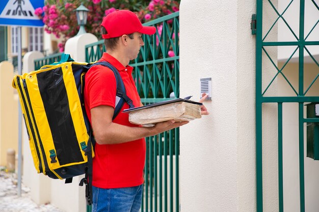 Geconcentreerde postbode aanbelt en houdt pakje en klembord vast. Knappe koerier in rode uniform draagtas, staat buiten en levert bestelling. Bezorgservice en postconcept