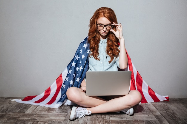 Geconcentreerde roodharige jonge dame met de vlag van de vs met behulp van laptop