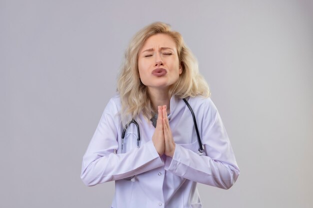 Geconcereerde jonge arts die stethoscoop in medische toga draagt die bidgebaar op isolatie witte muur toont
