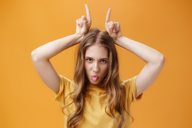 Gedurfde en coole emotionele aantrekkelijke vrouwelijke student met golvend blond haar en kleine littekens die opstandige tong tonen die van onder het voorhoofd kijken en wijsvingers op het hoofd houden die koppig zijn. Ruimte kopiëren