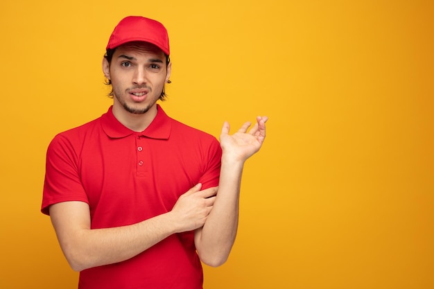 geen idee, jonge bezorger, gekleed in uniform en pet, kijkend naar de camera, houdend hand op arm, wijzend naar de andere kant met een andere hand geïsoleerd op gele achtergrond met kopieerruimte