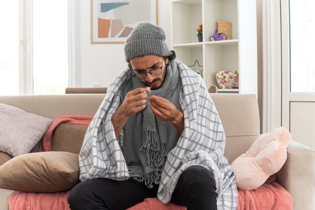 Geen idee, jonge, zieke man met optische bril gewikkeld in plaid met sjaal om zijn nek, wintermuts dragend en kijkend naar medische ampul en spuit zittend op de bank in de woonkamer