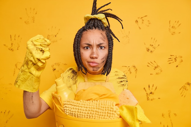 Gratis foto geërgerd afro-amerikaanse vrouw balt vuist kijkt boos naar camera poses vuil in de buurt van mand met wasgoed heeft dreadlocks geïsoleerd over gele muur gekamd
