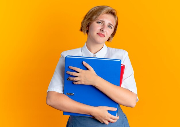 Geërgerd blond Russisch meisje houdt bestandsmappen geïsoleerd op een oranje achtergrond met kopie ruimte