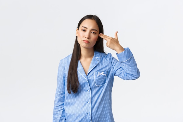Geërgerd en gehinderd Aziatisch meisje in blauwe pyjama dat met tegenzin kijkt, zichzelf neerschiet met een pistoolgebaar omdat ze het zat is, moe van het horen of zien van iets saais of doms, witte achtergrond.
