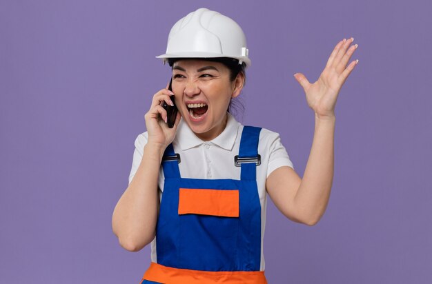 Geërgerd jong Aziatisch bouwmeisje met witte veiligheidshelm die tegen iemand aan de telefoon schreeuwt die naar de zijkant kijkt