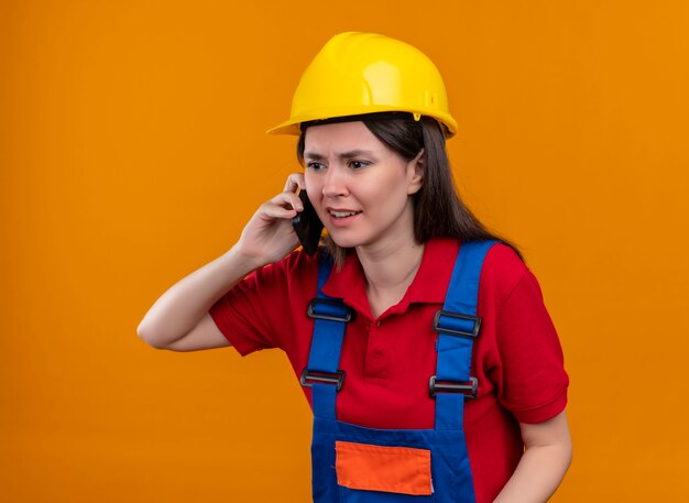 Geërgerd jong bouwersmeisje die op telefoon op geïsoleerde oranje achtergrond met exemplaarruimte spreken