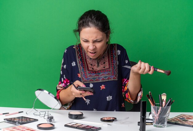 Geërgerd jong brunette meisje zittend aan tafel met make-up tools met make-up kwasten en kijken naar telefoon geïsoleerd op groene muur met kopie ruimte