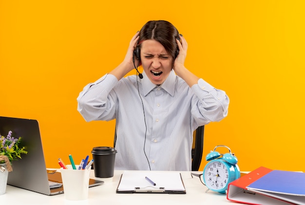 Geërgerd jong call centreermeisje die hoofdtelefoonszitting bij bureau dragen die haar hoofd met gesloten ogen houden die op oranje worden geïsoleerd