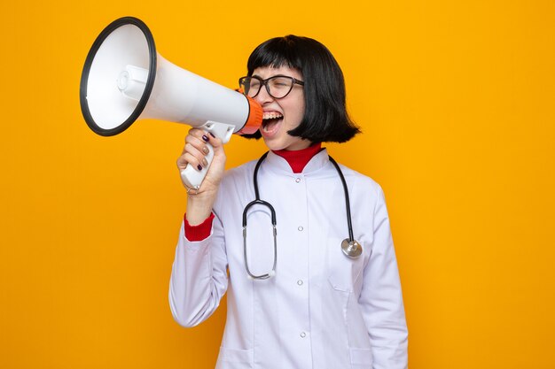 Geërgerd jong, vrij kaukasisch meisje met optische bril in doktersuniform met stethoscoop die in de luidspreker schreeuwt