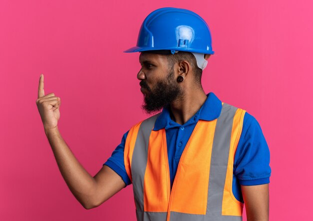 Geërgerd jonge Afro-Amerikaanse bouwer man in uniform met veiligheidshelm kijken naar zijn vinger geïsoleerd op roze achtergrond met kopie ruimte