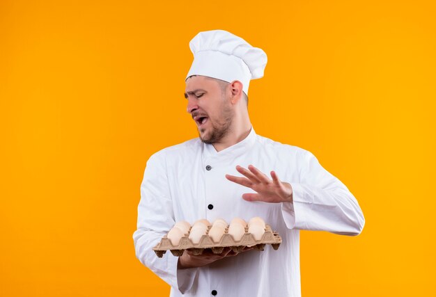 Geërgerd jonge knappe kok in uniform van de chef-kok met doos eieren gebaren nee geïsoleerd op oranje muur